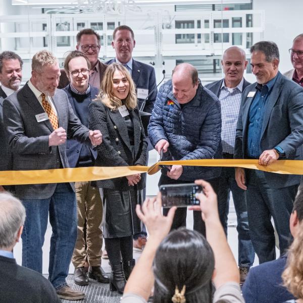 Man uses a large scissors to cut a golden ribbon with crowd gathering around him