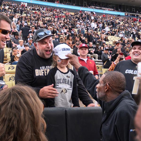 From the field, Coach Sanders reaches to put his baseball cap on the head a child. Witnesses to the interaction look on in amazement.