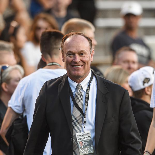 Rick George smiles, his gaze toward the camera, with people in the background in a stadium-like setting.