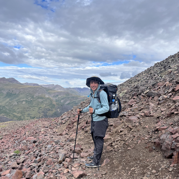 A man wearing a blue jacket and bucket hat smiles and hikes a rocky mountain.