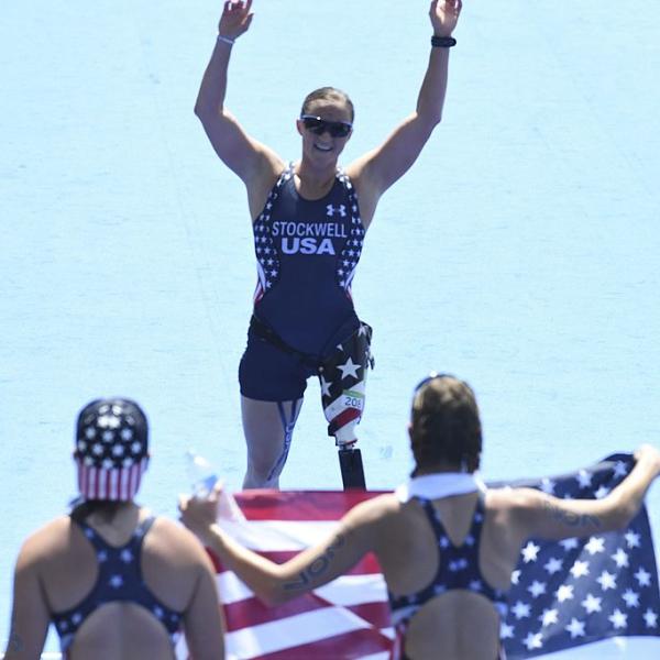 Melissa Stockwell, in stars-and-stripes track wear, with arms extended above her head, faces two people holding an American flag. (Wikimedia Commons/Tânia Rêgo/Agência Brasil)