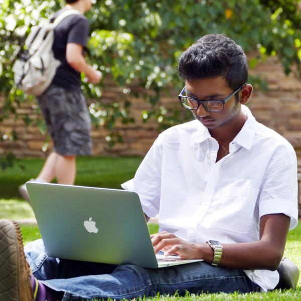 Student working on laptop, sitting in grass on campus