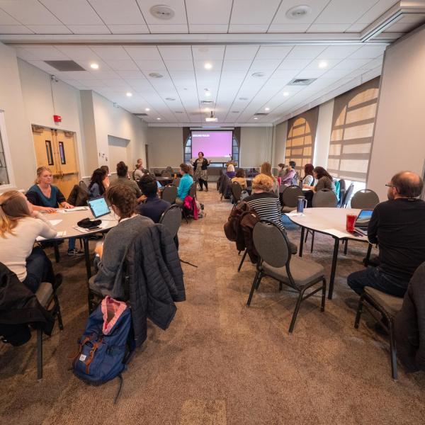 People sit at round tables and listen to a presentation.