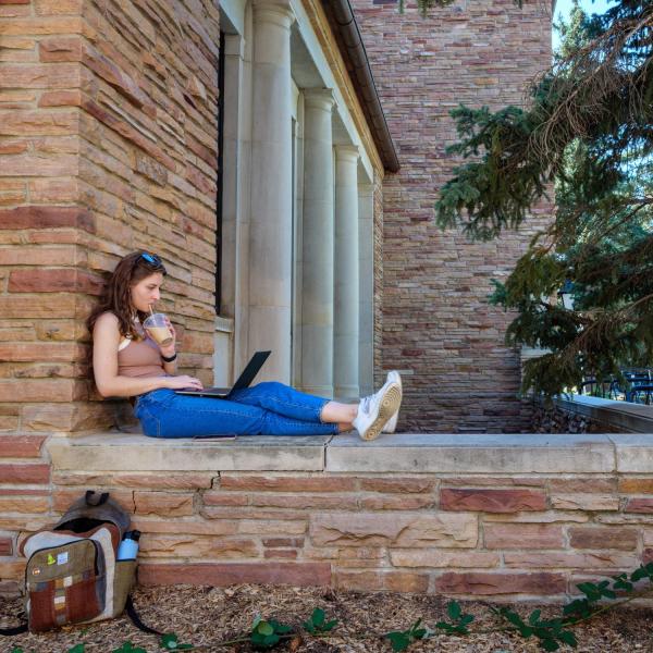 student sitting outside while working on laptop and drinking an ice coffee
