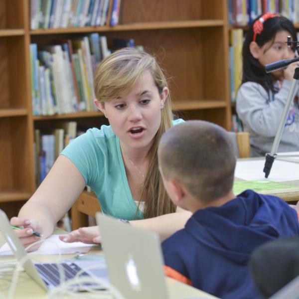 A college student gestures as she sits at a table and works with a young student in what appears to be a library setting.