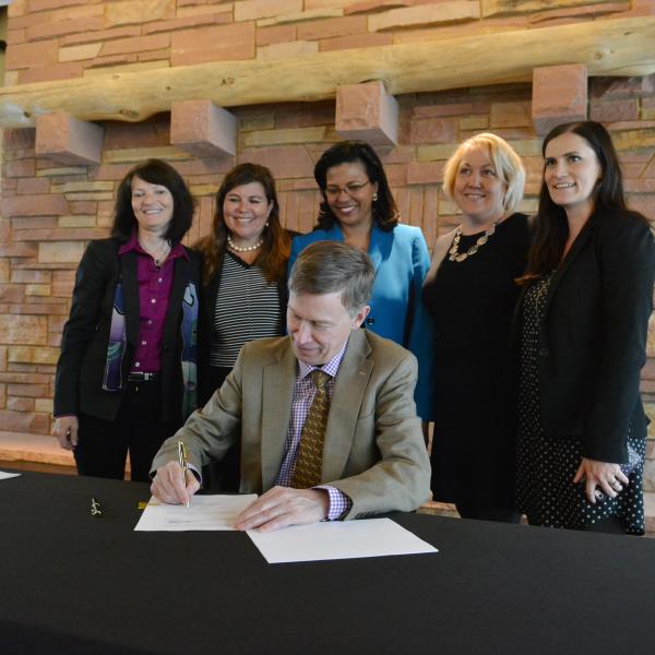 Gov. John Hickenlooper signs bill with people looking on.