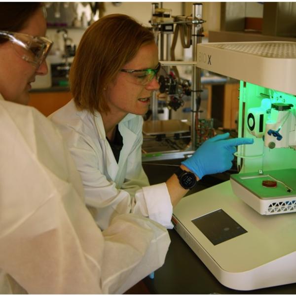 Two people in white lab coats, wearing safety goggles, work near lab equipment lit by a green light.