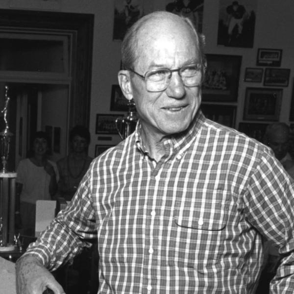 A black and white photo of Justice Byron White in a casual button down shirt with some trophies behind him.