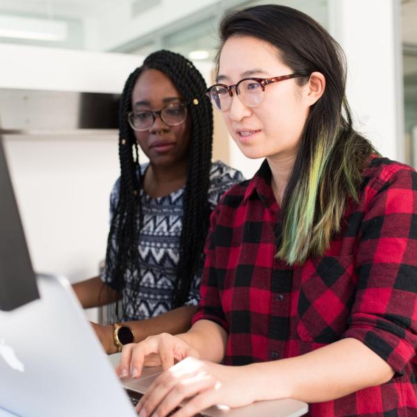 Two women working on a laptop computer at a standing desk.