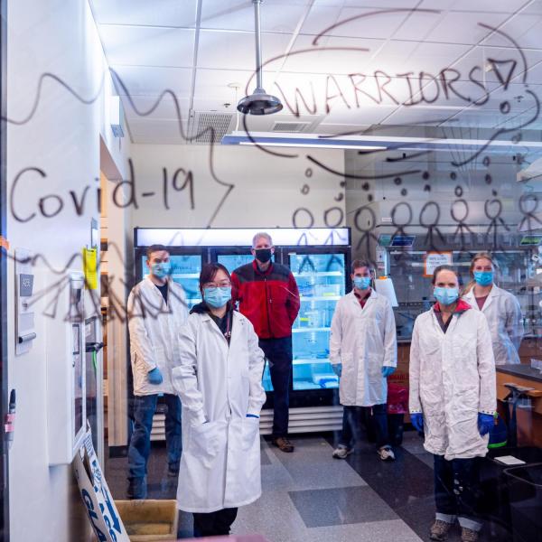 Researchers pose in their lab behind glass with the words "COVID Warriors" written on it.