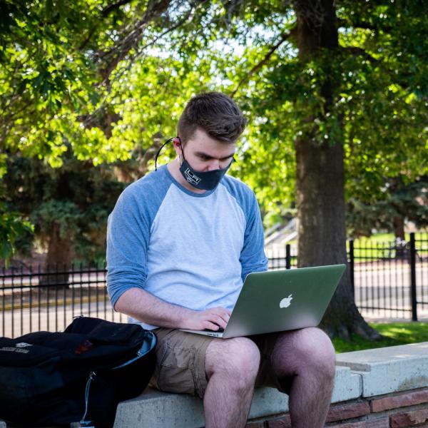 Student in mask works on laptop outside