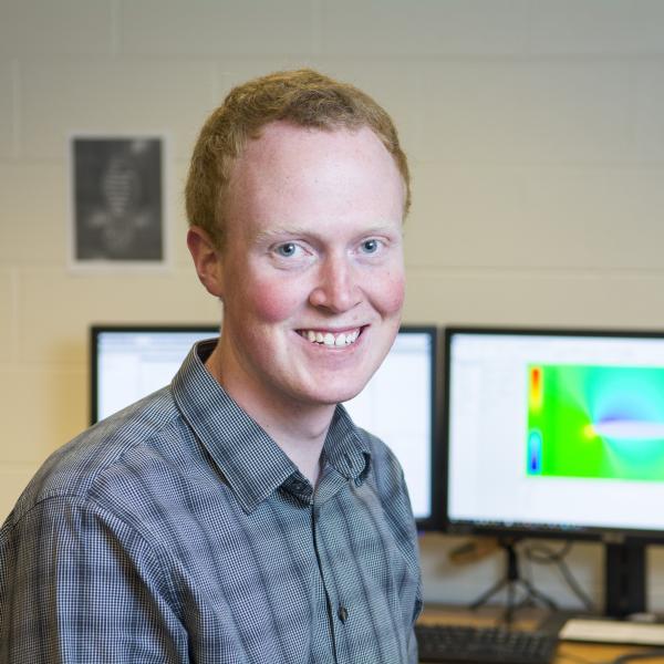 Portrait of CU student Matthew Hurst in front of monitors displaying his computer modeling graphics