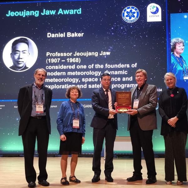 Dan Baker holds a plaque and poses for a group photo on a stage.