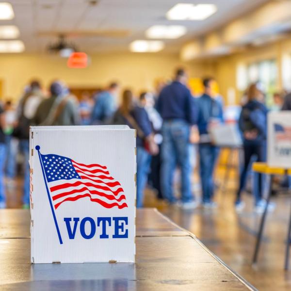 Sign with American flag and the word "Vote" sits out on a table while people mill around in the background