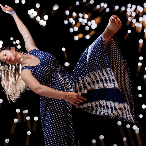 In a horizontal dance position, Erika Randall extends her left leg with a pointed toe, and reaches back with her left arm, round golf-ball size lights glimmering around her with a black background.