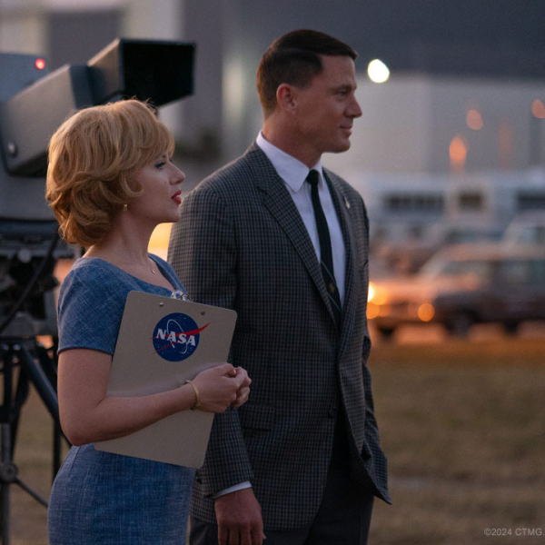 Man and women stand side-by-side with camera in the background. Woman carries a clipboard labeled "NASA"