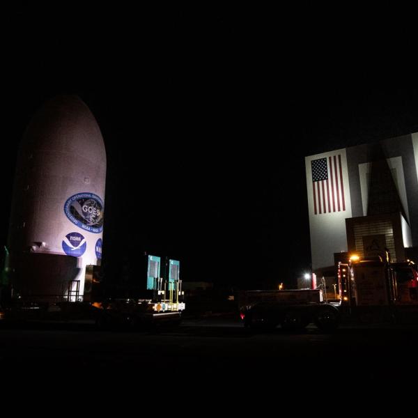 Satellite labeled "GOES" sits next to a large hangar labeled "NASA" at night time