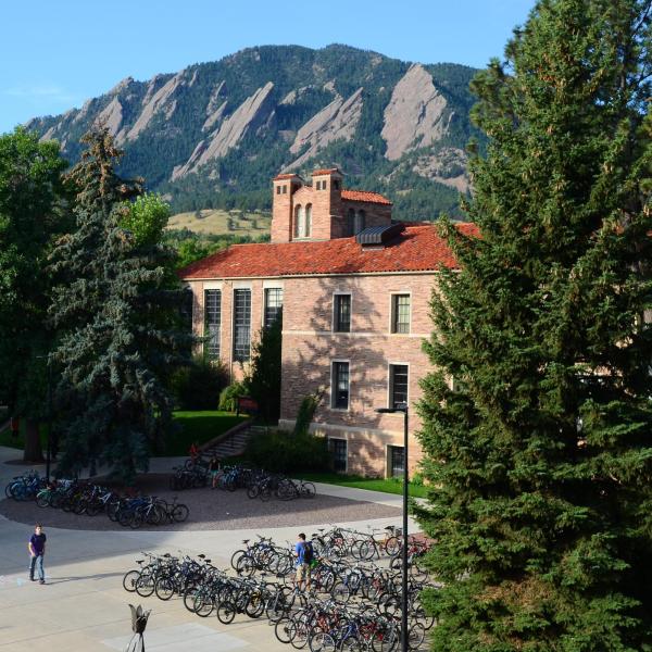 The CU Boulder Hellems building is brightly lit by the sun, with evergreen trees flanking it and the Flatirons and a blue sky in the background.