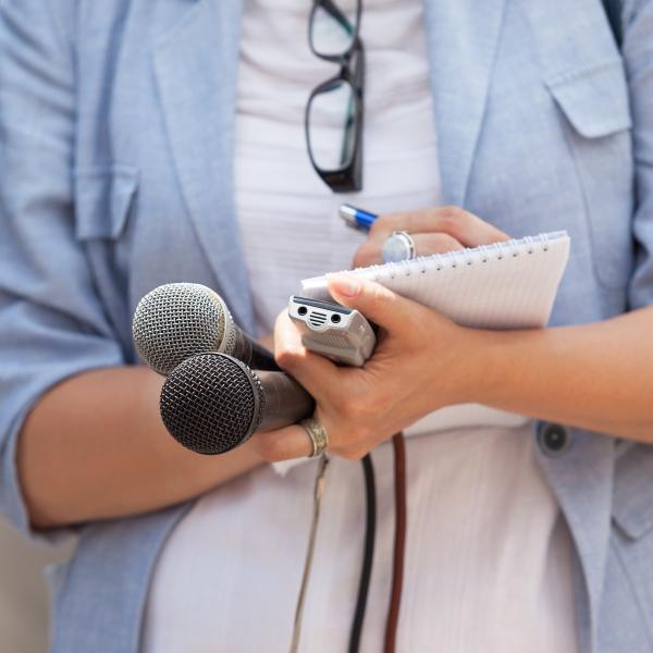 In a stock image, a journalist stands with two microphones, a reporter's notebook and pen.