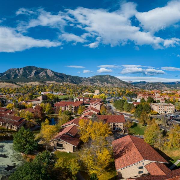 An aerial view of part of the CU Boulder campus shows colorful fall foliage, the Flatirons in the distance and a bright blue sky with light clouds.