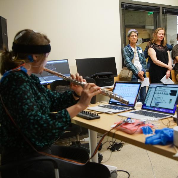 Grace Leslie plays the flute in front oof computer screens while a small group of people look on