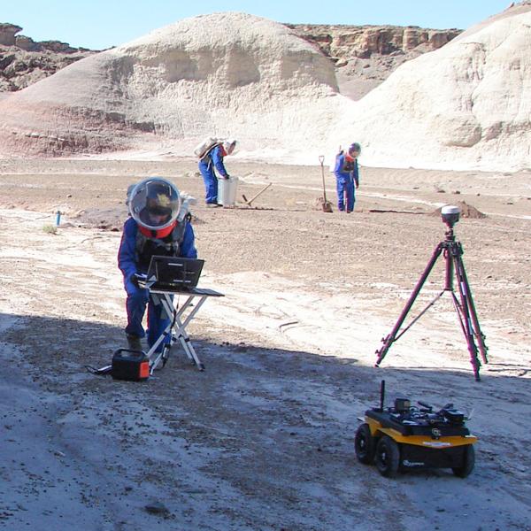 Three people in spacesuit-like gear appear to work with equipment in a dry hilly outdoor space.