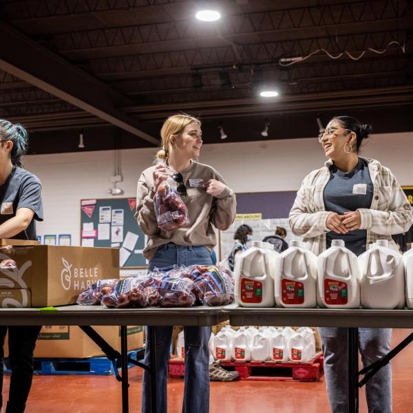 People stand at a table with bags of produce and gallon jugs of milk.