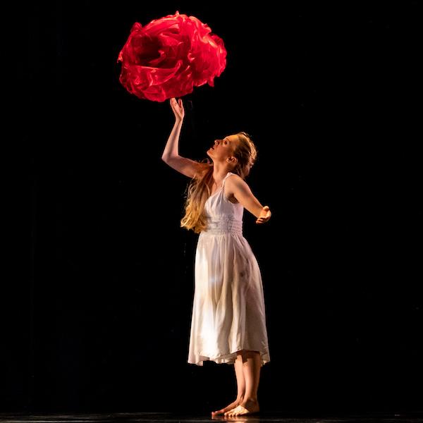 A dancer in a white flowing dress on stage with large red flower shapes.