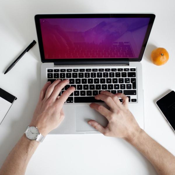 a person working on a laptop with a notebook, pen, phone and an orange nearby