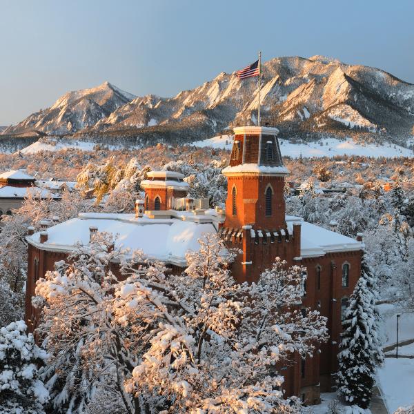 Snowy Old Main tower with American flag overlooking the flatirons