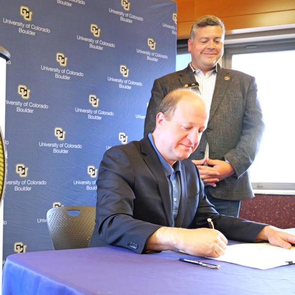 Man sits at table to sign document while another man looks on. A podium bearing the seal of the State of Colorado is in the foreground