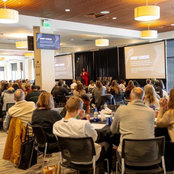 Numerous people sit at large round tables in a conference space, turning their attention to a speaker holding a microphone and presenting from a lecturn.