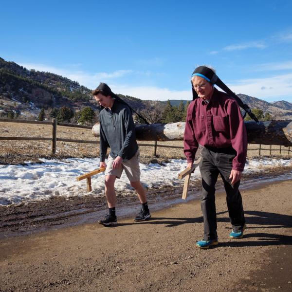 James Wilson and Rodger Kram carry a log from their heads using tumplines with the Boulder Foothills in the background