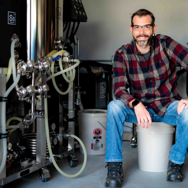 Man in flannel shirt wearing safety glasses sits next to equipment with lots of tubes