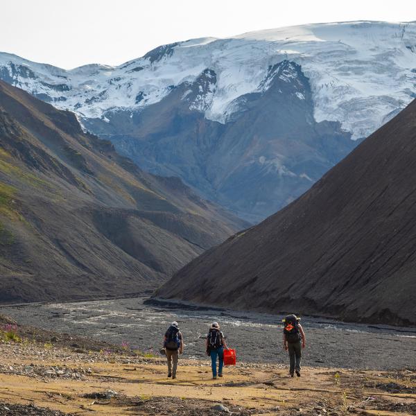 People walking toward a snow-covered mountain through a dry river bed of rocks.