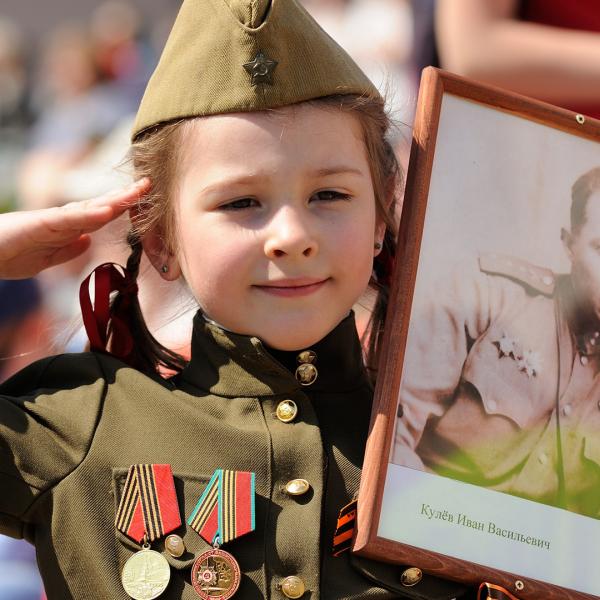 Young Russian girl wearing military uniform and medals carrying soldier's portrait during anniversary celebration of Victory Day