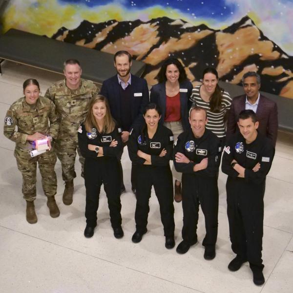 10 people, four wearing black flight suits and tow wearing camouflage uniforms, pose for a photo as seen from above