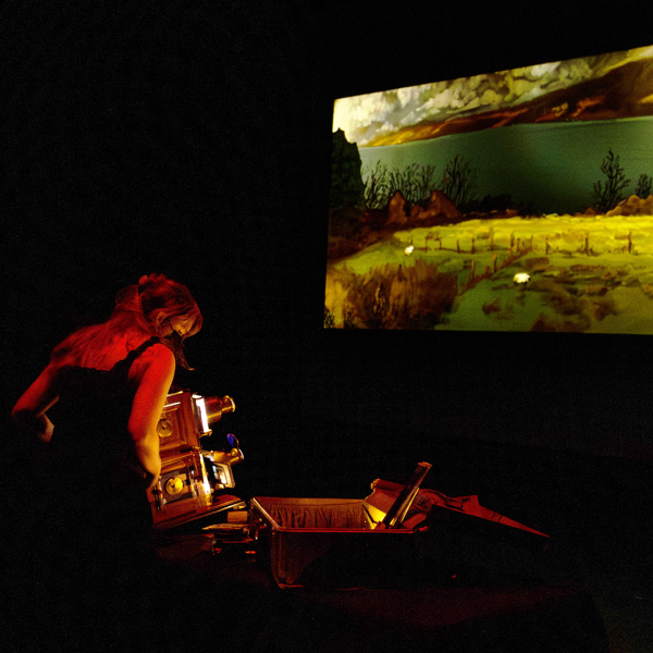 A person stands at a console, looking down. The room is dark and shows a split-screen projection of a bolder in water, and a green landscape.