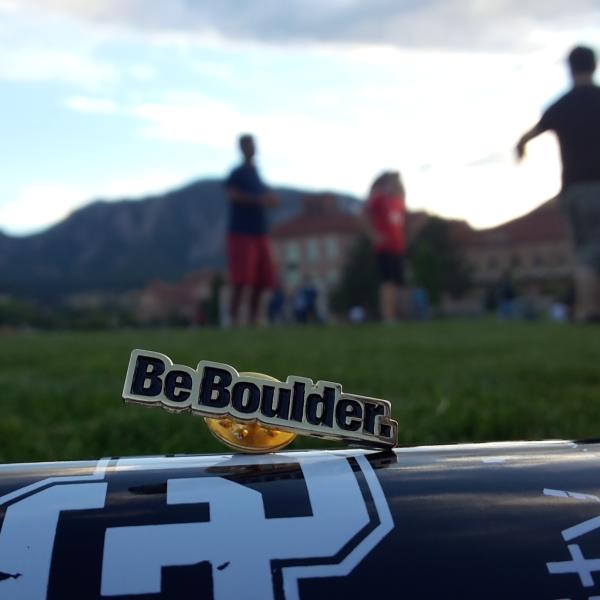 A Be Boulder pin sits atop poster paper on a field on campus with people in the background.