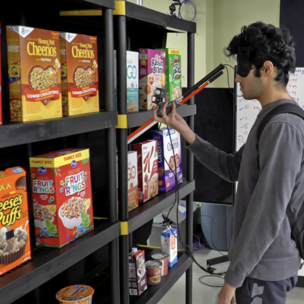 Man wearing blindfold holds up a camera on a walking stick to scan a shelf containing several different brands of cereal.