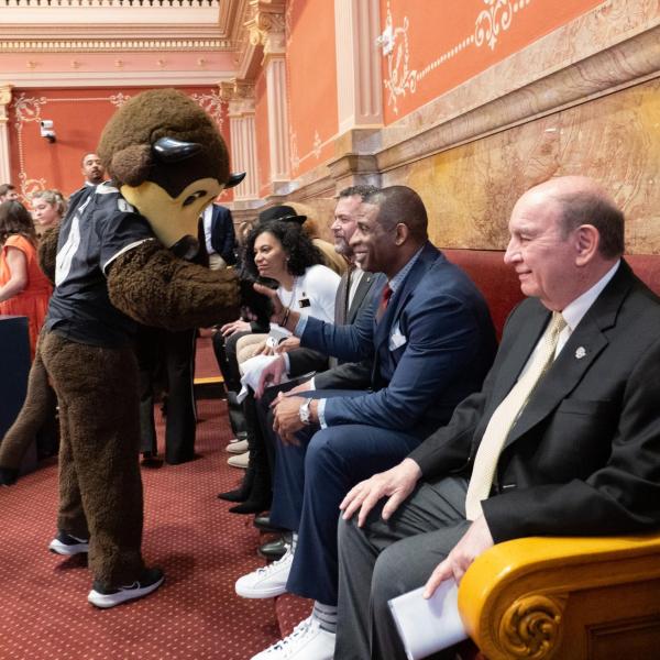 Deion Sanders, sitting next to Chancellor Phil DiStefano, greets Chip the buffalo mascot before being honored in the Senate Chamber