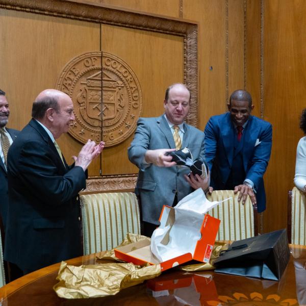 Gov. Jared Polis opens a box of CU Buffalo-branded shoes as Todd Saliman, Phil DiStefano, Deion Sanders and Wanda James look on