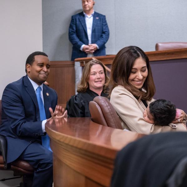 U.S. Representative Joe Neguse and Colorado Supreme Court justice Melissa Hart, center, and Andrea Neguse holding a baby listen to Governor Jared Polis speak. Photo by Glenn Asakawa.
