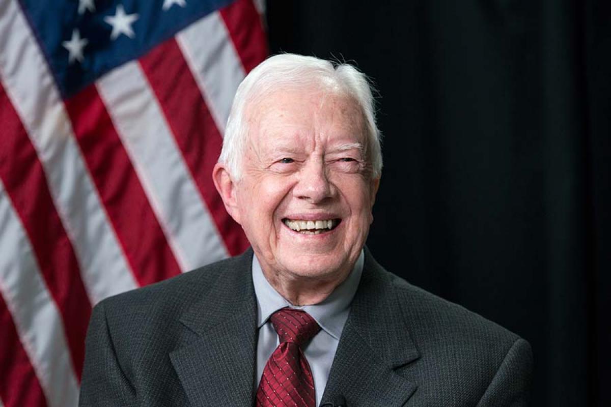 The late President Jimmy Carter in suit and red tie with an American flag in the background