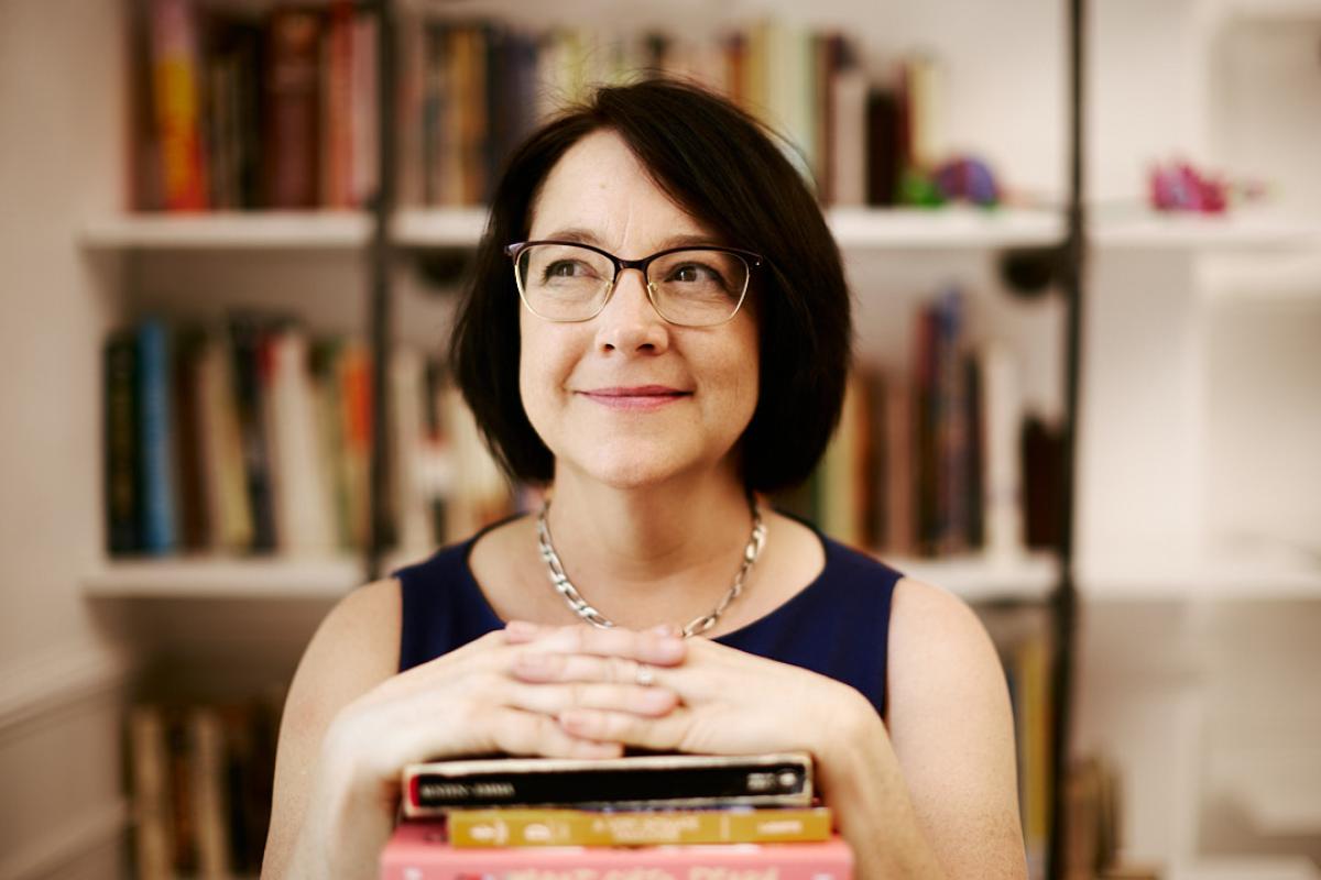 Woman crosses hands over a pile of books with a bookshelf in the backgroundbra