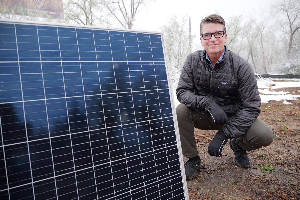 Man kneels on the ground next to a large solar panel with snow in the background