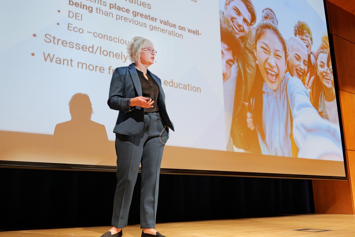 A person in a suit, holding a remote in her hand stands on a stage with a screen and slide projection behind her showing bullet points and a picture of a group of people looking happy and excited.