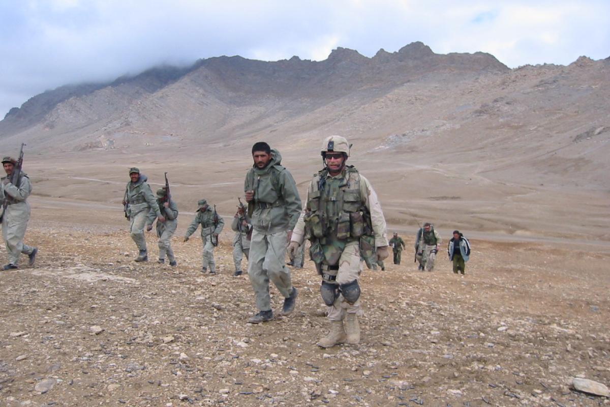 Mitch Utterback along with several other members of armed forces walk in what appears to be a dry climate, out in open space with mountains in the background.