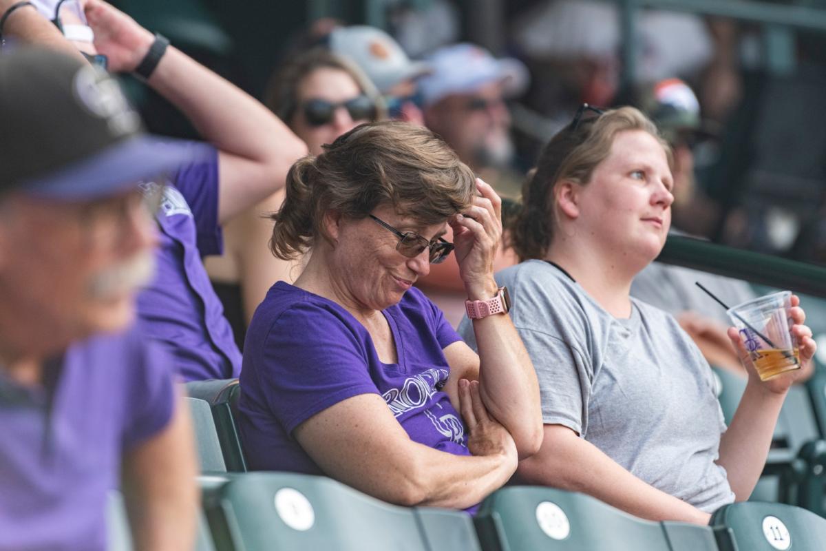 A person in a purple Rockies T-shirt sits in a stadium, one arm crossed and the other extended to scratch her head.