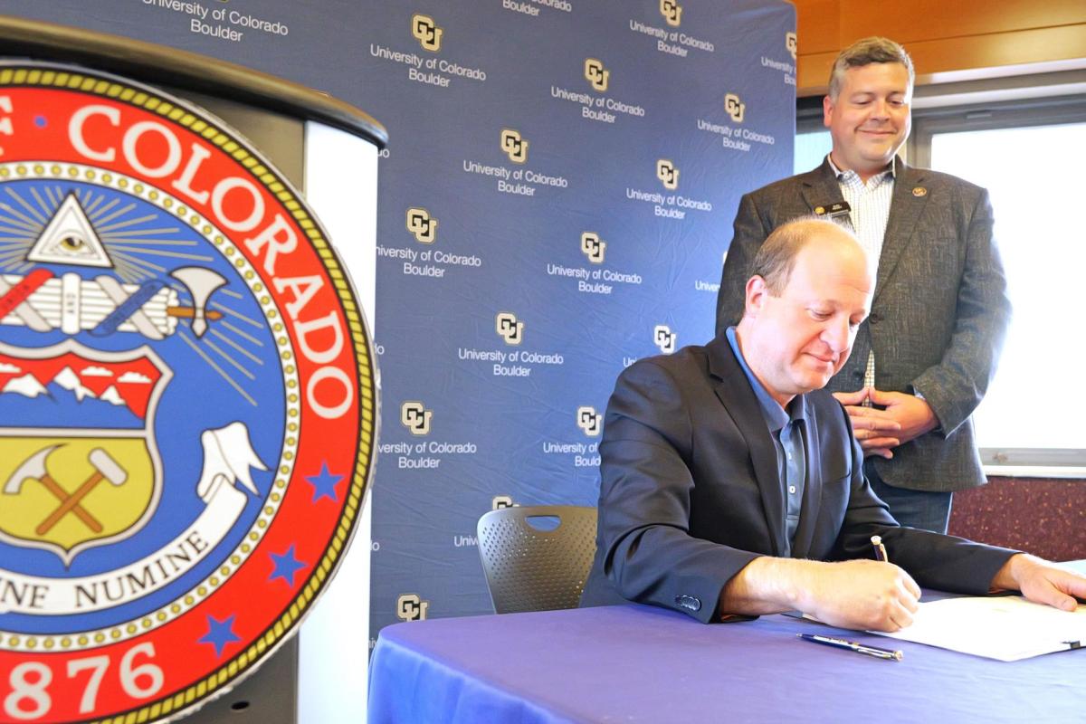 Man sits at table to sign document while another man looks on. A podium bearing the seal of the State of Colorado is in the foreground
